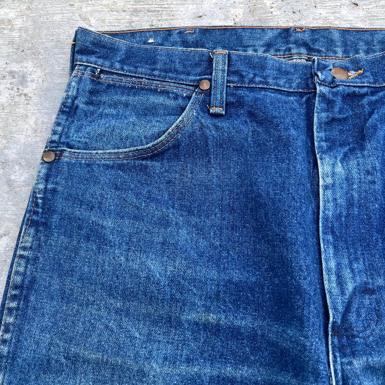 Pode incluir: Close-up de jeans azuis. Os jeans t&ecirc;m um design cl&aacute;ssico de cinco bolsos com fecho de bot&atilde;o e z&iacute;per. O tecido mostra sinais de desgaste, com desbotamento sutil e uma apar&ecirc;ncia texturizada.