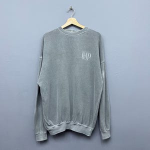 May include: A grey sweatshirt with long sleeves and the word "GAP" embroidered on the upper left chest. The garment is suspended from a wooden hanger. The background is a plain grey. The fabric has a faded appearance.
