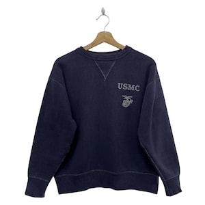 May include: Navy blue sweatshirt with the text "USMC" and a silver emblem. The crew neck sweatshirt is hanging on a wooden hanger. The sweatshirt has a relaxed fit and ribbed cuffs and hem.