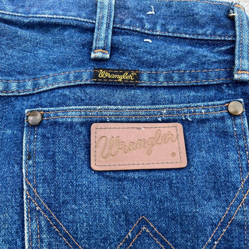 Pode incluir: Close-up do bolso traseiro de jeans azuis. Os jeans apresentam um patch de couro marrom com o logotipo "Wrangler" e uma pequena etiqueta "Wrangler" acima do bolso. A costura &eacute; de cor laranja contrastante.
