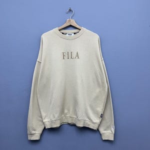 May include: A beige crewneck sweatshirt featuring the FILA logo embroidered in a matching tan thread on the chest. The garment has long sleeves, ribbed cuffs, and a ribbed hem, with a branded interior neck label and a small logo tag on the side.