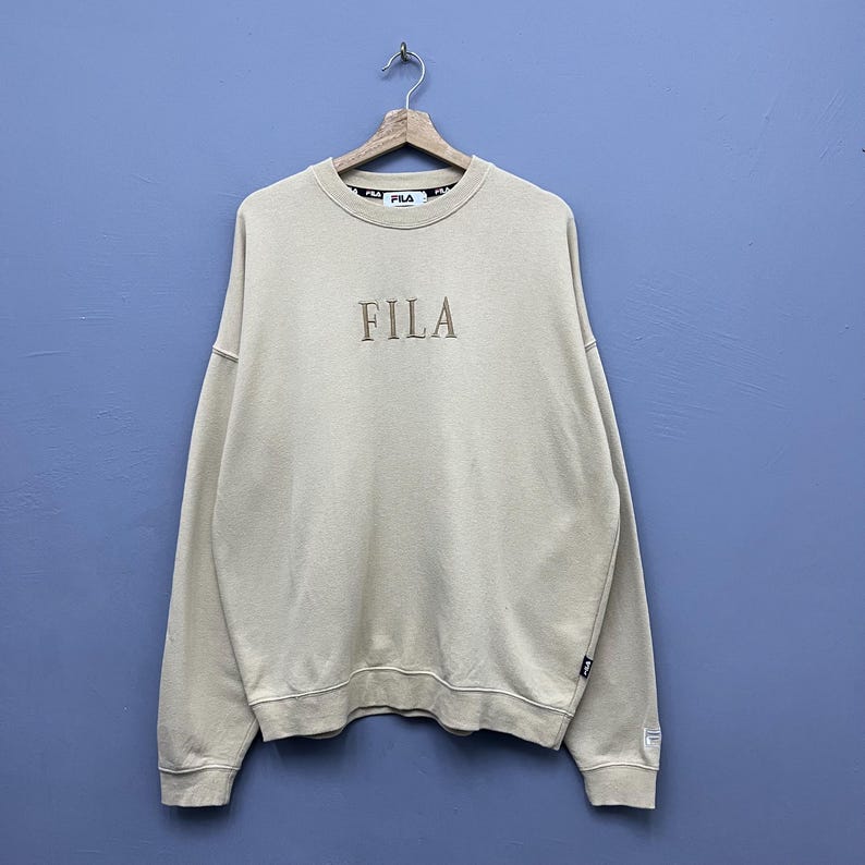 May include: A beige crewneck sweatshirt featuring the FILA logo embroidered in a matching tan thread on the chest. The garment has long sleeves, ribbed cuffs, and a ribbed hem, with a branded interior neck label and a small logo tag on the side.