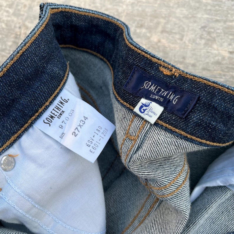 May include: Close-up of dark wash denim jeans with visible stitching and tags. The tags read "SOMETHING EDWIN" and include size information: 27x34. The jeans have a classic five-pocket design.