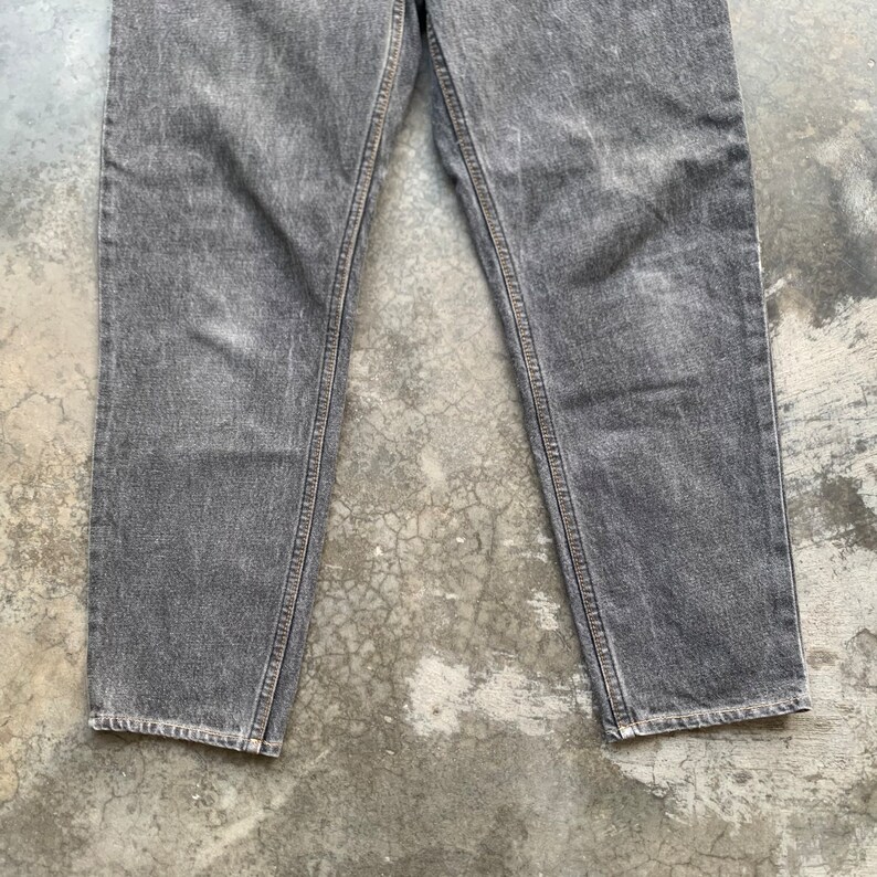 May include: Close-up of gray denim jeans. The jeans have a classic five-pocket design, with visible stitching along the seams and a faded appearance. The legs are straight cut, and the fabric appears to be a medium weight.