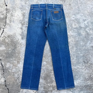 May include: Back view of blue denim jeans with classic five-pocket styling. The jeans feature a brown leather patch on the back and a Wrangler label. The denim has a faded appearance, suggesting a vintage or worn look.