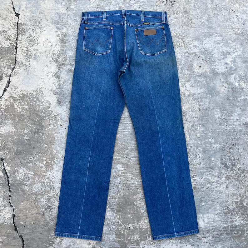 May include: Back view of blue denim jeans with classic five-pocket styling. The jeans feature a brown leather patch on the back and a Wrangler label. The denim has a faded appearance, suggesting a vintage or worn look.