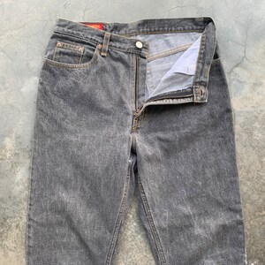 May include: A pair of gray denim jeans with a button and zipper closure. The jeans have a classic five-pocket design and a faded wash. The waistband features a red tag. The jeans are laid flat on a concrete surface.