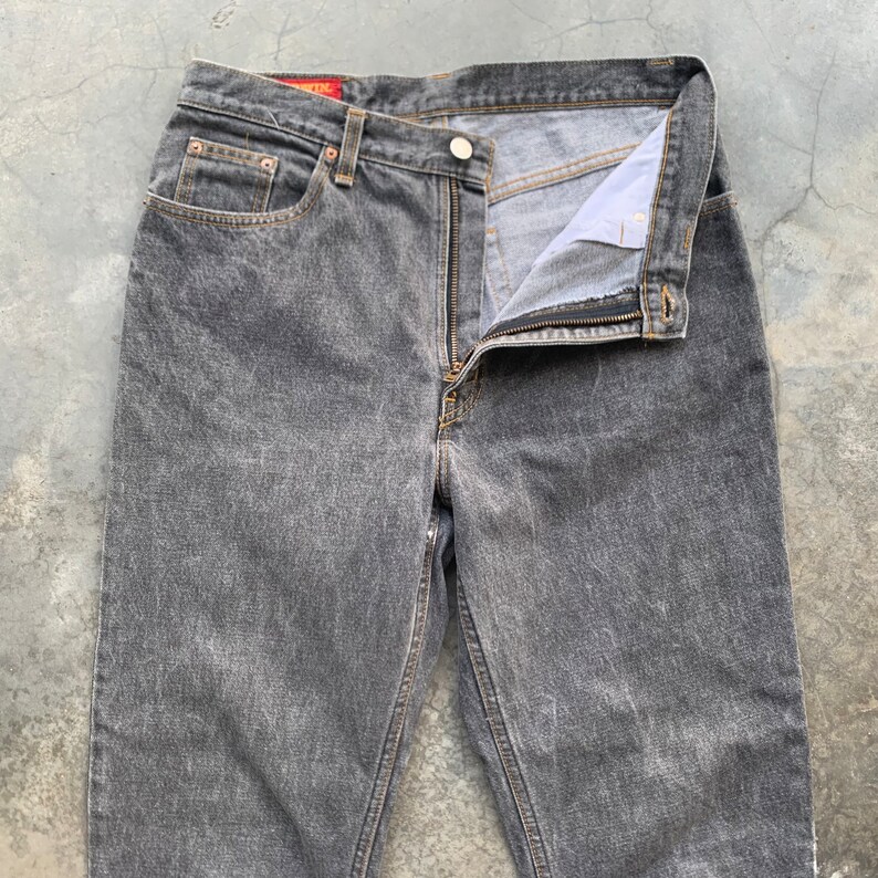 May include: A pair of gray denim jeans with a button and zipper closure. The jeans have a classic five-pocket design and a faded wash. The waistband features a red tag. The jeans are laid flat on a concrete surface.