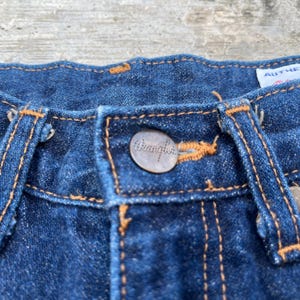 K&ouml;nnte beinhalten: Nahaufnahme einer blauen Jeans. Die Jeans hat einen Metallknopf mit dem Markennamen "Wrangler" und orangefarbenen N&auml;hten. Das Bild zeigt die Details des Knopfes, der N&auml;hte und der Stoffstruktur.