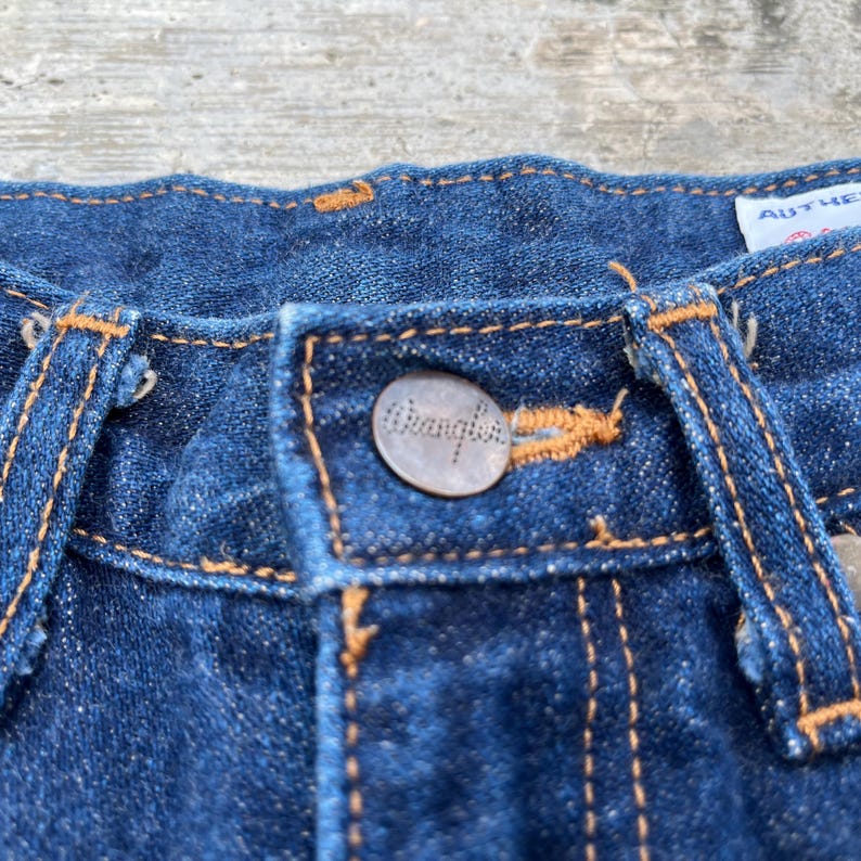 K&ouml;nnte beinhalten: Nahaufnahme einer blauen Jeans. Die Jeans hat einen Metallknopf mit dem Markennamen "Wrangler" und orangefarbenen N&auml;hten. Das Bild zeigt die Details des Knopfes, der N&auml;hte und der Stoffstruktur.