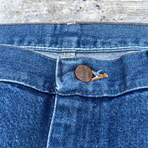 May include: Close-up of blue denim jeans with a copper-colored button. The button is embossed with a logo. The jeans have a classic five-pocket design and are made of durable denim fabric. The stitching is in a contrasting color.