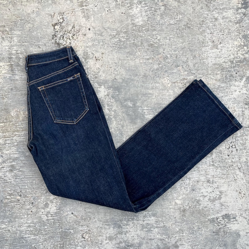 May include: Dark wash denim jeans with a classic five-pocket design. The jeans have a straight leg cut and a button and zipper closure. The back pocket features a small brand label. The jeans are laid flat on a gray surface.