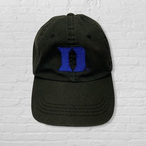 May include: A black baseball cap featuring a prominent blue embroidered letter D on the front panel. The cap has a structured crown, a curved brim with visible stitching, and a small registered trademark symbol next to the blue letter design.