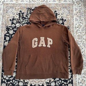 Könnte beinhalten: Ein brauner Kapuzenpullover mit dem Wort "GAP" in cremefarbenen Blockbuchstaben auf der Brust. Der Pullover hat eine Kängurutasche und eine Kapuze mit Kordelzug. Das Kleidungsstück ist aus weichem Material.