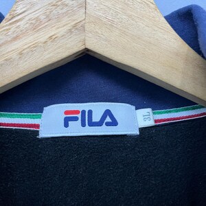 Puede incluir: Primer plano de una prenda azul marino con una etiqueta blanca con el logotipo de FILA en azul y rojo. La prenda tiene una franja tricolor y una etiqueta de talla que dice "3L". La prenda est&aacute; colgada en una percha de madera.