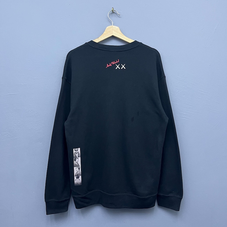 May include: A black long-sleeve crewneck sweatshirt featuring a red Andy Warhol signature and white double X graphic on the upper chest. A vertical strip of black and white photographic film-style images is printed on the lower right side.