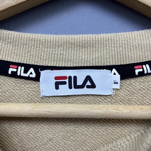 May include: A beige sweatshirt featuring a ribbed crew neckline with a black interior trim printed with the white and red Fila logo. A white rectangular brand label is stitched at the center, displaying the Fila logo and a small tag with the letter F.
