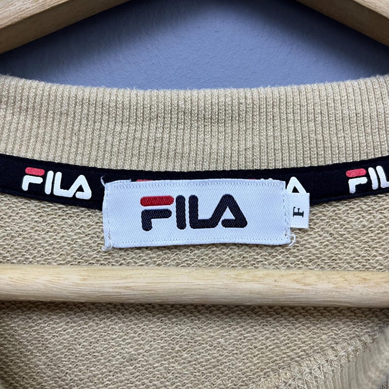 May include: A beige sweatshirt featuring a ribbed crew neckline with a black interior trim printed with the white and red Fila logo. A white rectangular brand label is stitched at the center, displaying the Fila logo and a small tag with the letter F.