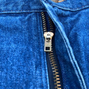 Pode incluir: Close-up de um tecido denim azul com um z&iacute;per de lat&atilde;o. O z&iacute;per est&aacute; parcialmente fechado, revelando os dentes e o puxador de metal. O denim tem uma apar&ecirc;ncia texturizada.