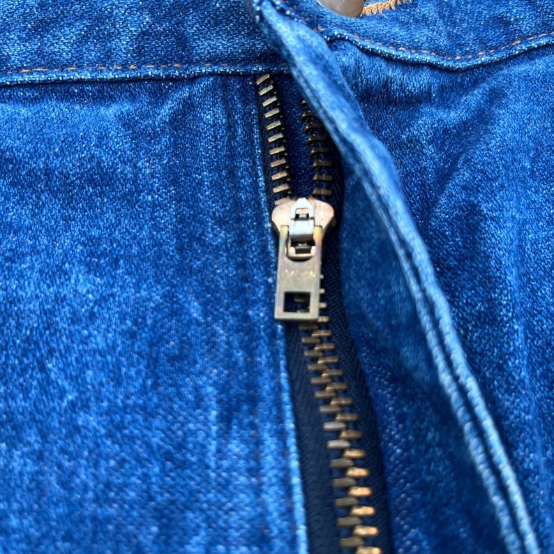 Pode incluir: Close-up de um tecido denim azul com um z&iacute;per de lat&atilde;o. O z&iacute;per est&aacute; parcialmente fechado, revelando os dentes e o puxador de metal. O denim tem uma apar&ecirc;ncia texturizada.