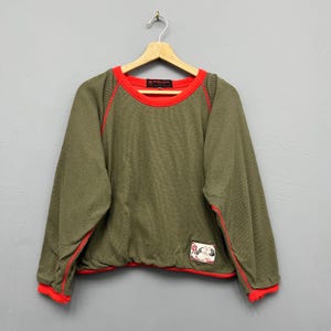 May include: An olive green long-sleeve thermal shirt featuring a bright red crew neckline, red raglan sleeve piping, and red cuffs. The hem is elasticized with a red trim, and a small rectangular patch with a graphic illustration is on the lower left.