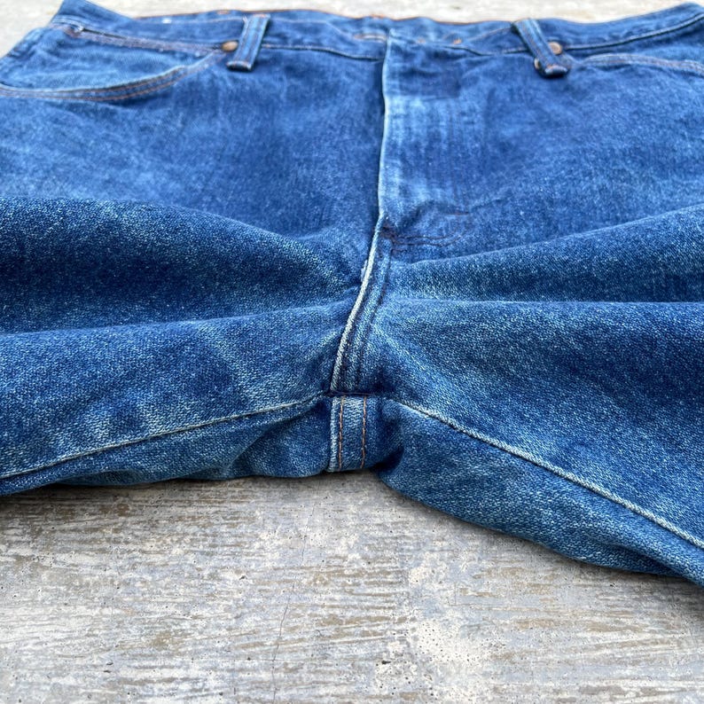 Pode incluir: Close-up de jeans de ganga azul. Os jeans t&ecirc;m um design cl&aacute;ssico de cinco bolsos, com fecho de bot&atilde;o e z&iacute;per. A ganga tem um aspeto desbotado e usado, com costuras e rebites vis&iacute;veis.
