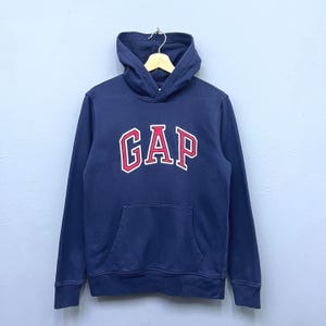 May include: Navy blue hooded sweatshirt with the word "GAP" in red letters with a white outline across the chest. The sweatshirt has a kangaroo pocket and a drawstring hood. The garment is hanging on a wooden hanger.