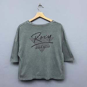 Puede incluir: Camiseta verde oliva de manga corta con el logo de Roxy Surf Club. El logotipo negro presenta el nombre Roxy en una escritura estilizada sobre las palabras "surf club" dentro de un triángulo.