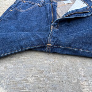 K&ouml;nnte beinhalten: Nahaufnahme einer blauen Jeans mit teilweise ge&ouml;ffnetem Rei&szlig;verschluss. Die Jeans hat einen klassischen F&uuml;nf-Taschen-Stil mit orangefarbenen N&auml;hten. Der Stoff weist eine strukturierte Optik auf, und die Jeans liegt auf einer grauen Oberfl&auml;che.