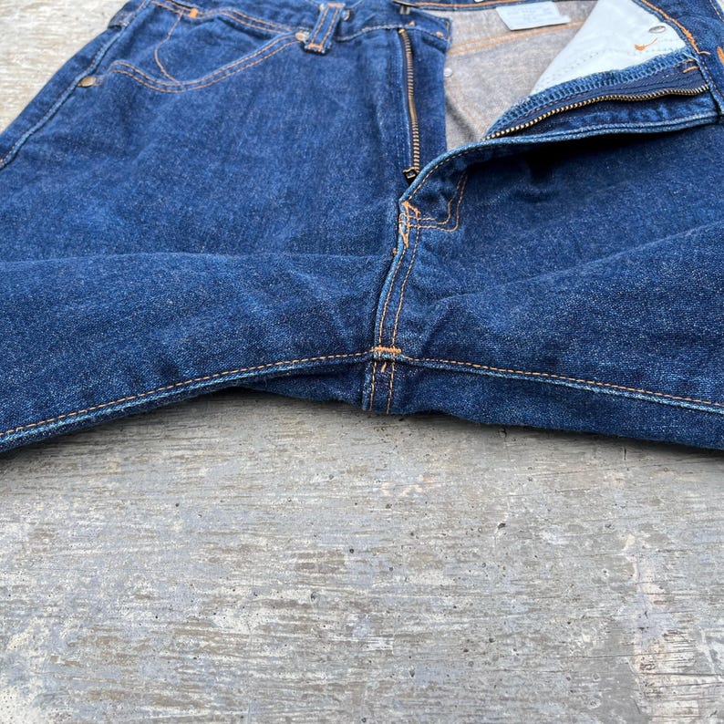 K&ouml;nnte beinhalten: Nahaufnahme einer blauen Jeans mit teilweise ge&ouml;ffnetem Rei&szlig;verschluss. Die Jeans hat einen klassischen F&uuml;nf-Taschen-Stil mit orangefarbenen N&auml;hten. Der Stoff weist eine strukturierte Optik auf, und die Jeans liegt auf einer grauen Oberfl&auml;che.