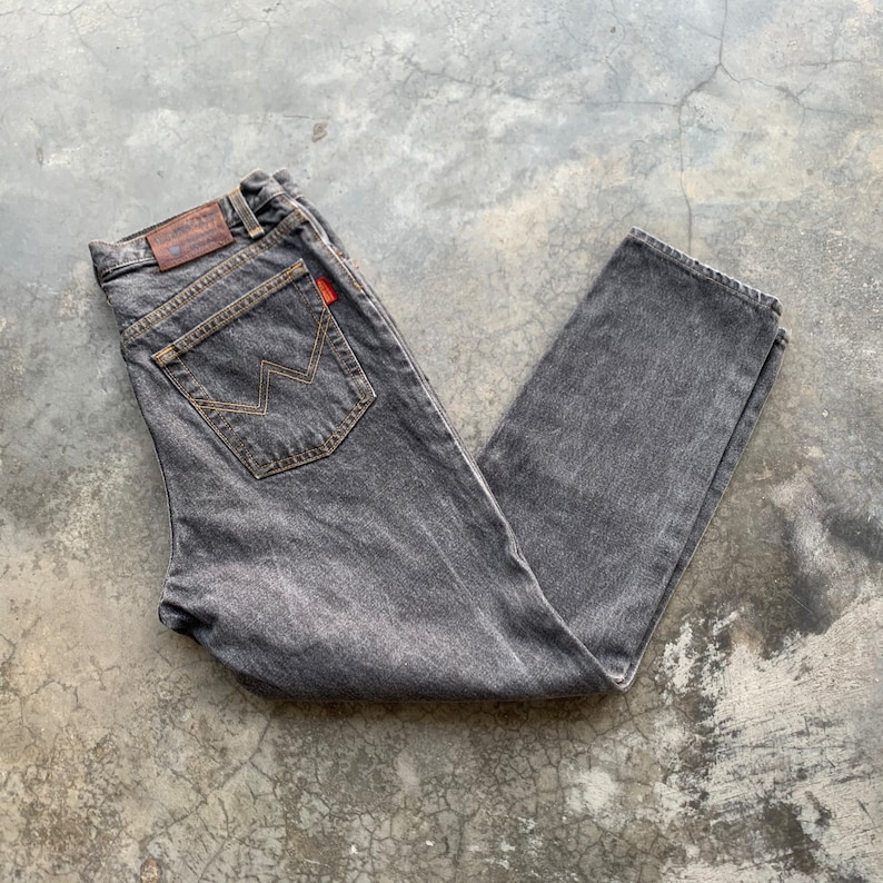 May include: A pair of faded black denim jeans. The jeans have a classic five-pocket design, with a brown leather patch on the back waistband and a red tag on the back pocket. The stitching is a contrasting tan color.