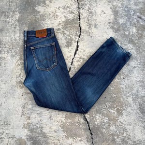 May include: Dark blue denim jeans with a classic five-pocket design. The jeans feature a brown leather patch with the brand name "EDWIN" on the back waistband. The jeans are laid flat on a concrete surface.