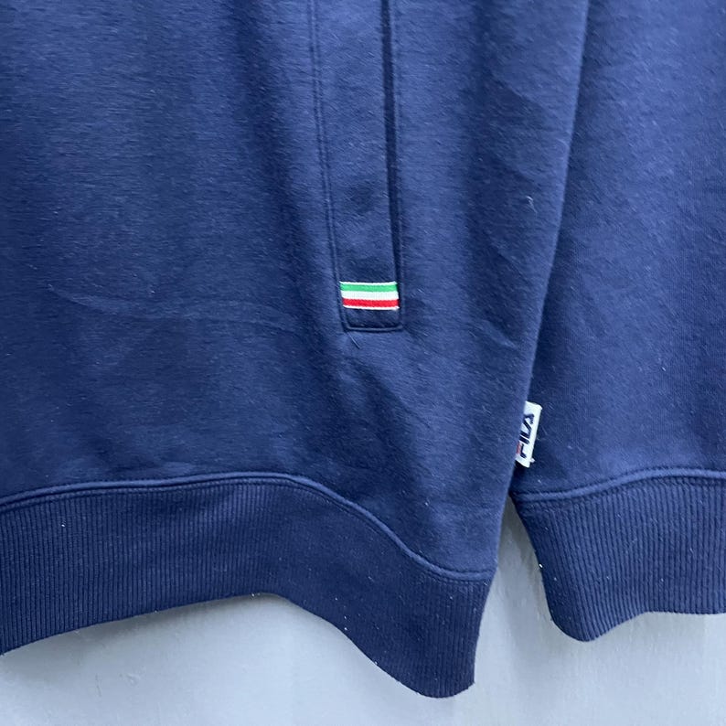 Puede incluir: Sudadera azul marino con un peque&ntilde;o bolsillo y un detalle de rayas tricolores en verde, blanco y rojo. El logotipo de la marca es visible en la parte inferior derecha. La sudadera es de un material suave.