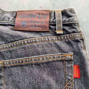 May include: Close-up of the back of a pair of faded black denim jeans. The jeans feature a brown leather patch with the brand name "EDWIN" and "MADE IN U.S.A." The stitching is a contrasting yellow, and a red tag with the brand name is visible.