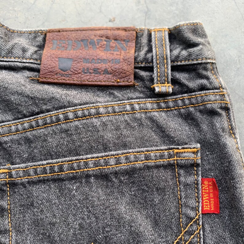 May include: Close-up of the back of a pair of faded black denim jeans. The jeans feature a brown leather patch with the brand name "EDWIN" and "MADE IN U.S.A." The stitching is a contrasting yellow, and a red tag with the brand name is visible.