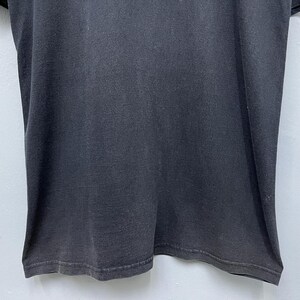 Op de afbeelding: Een close-up van een donkergrijs t-shirt. De stof lijkt licht versleten, met subtiele kleurvariaties. De zoom aan de onderkant is zichtbaar en het shirt heeft een eenvoudig, klassiek ontwerp.