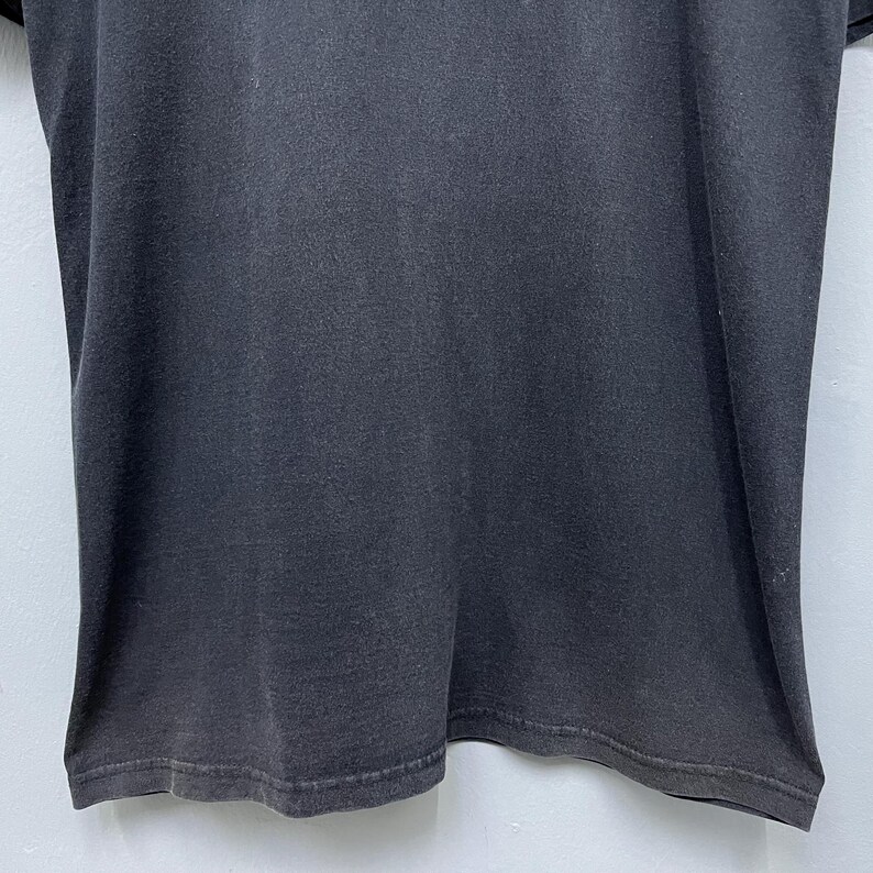 Op de afbeelding: Een close-up van een donkergrijs t-shirt. De stof lijkt licht versleten, met subtiele kleurvariaties. De zoom aan de onderkant is zichtbaar en het shirt heeft een eenvoudig, klassiek ontwerp.
