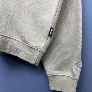May include: A beige long-sleeve sweatshirt featuring a ribbed hem and cuffs. A small navy blue tag with the white and red Fila logo is sewn into the side seam. An embroidered white Fila logo is visible on the lower portion of the sleeve.