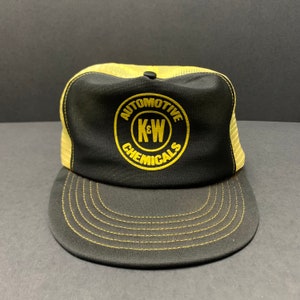 May include: Black and gold trucker hat featuring the text "AUTOMOTIVE CHEMICALS" and "K&W" in a circular design. The hat has a mesh back and a curved peak. A classic style.