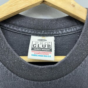 Op de afbeelding: Close-up van een donkergrijze t-shirtkraag met een wit label. Het label leest "PRO CLUB HEAVY WEIGHT" en bevat details over stof en assemblage. Het shirt is gemaakt in de VS.