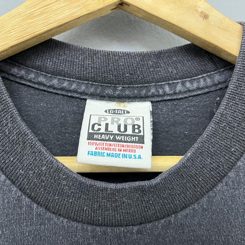 Op de afbeelding: Close-up van een donkergrijze t-shirtkraag met een wit label. Het label leest "PRO CLUB HEAVY WEIGHT" en bevat details over stof en assemblage. Het shirt is gemaakt in de VS.