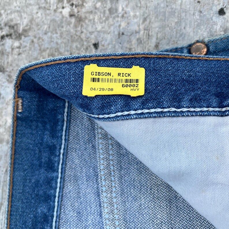 May include: Close-up of blue denim jeans with a yellow tag. The tag reads "GIBSON, RICK" and includes a barcode, the date "04/29/08", and the code "60002 HVY". The jeans have a light blue wash and visible stitching.