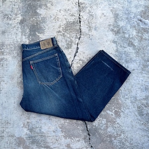 Könnte beinhalten: Dunkelblaue Jeans mit klassischem Fünf-Taschen-Design. Die Jeans haben einen Lederaufnäher auf der Rückseite und ein rotes Etikett auf der Gesäßtasche. Die Jeans liegen flach auf einer strukturierten, grauen Oberfläche.