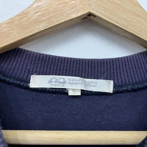 May include: Close-up of a navy blue sweatshirt with a ribbed neckline. The label reads "Courreges Sport Futur Paris" with a logo and the number "5". The sweatshirt is hanging on a wooden hanger.