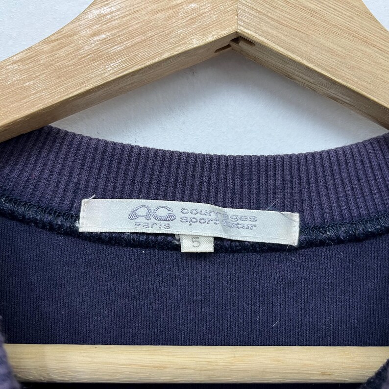 May include: Close-up of a navy blue sweatshirt with a ribbed neckline. The label reads "Courreges Sport Futur Paris" with a logo and the number "5". The sweatshirt is hanging on a wooden hanger.