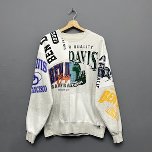 Puede incluir: Una sudadera de cuello redondo gris claro con un diseño de patchwork que presenta varios logotipos, textos y gráficos de la marca Ben Davis en negro, verde, azul, naranja y amarillo. La prenda incluye una ilustración de un globo y el texto San Francisco.