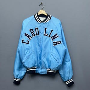 May include: Light blue satin bomber jacket with the word "CAROLINA" arched across the chest in navy blue lettering. The jacket has a navy blue and white striped collar, cuffs, and hem. Features two front pockets and a button-up closure.