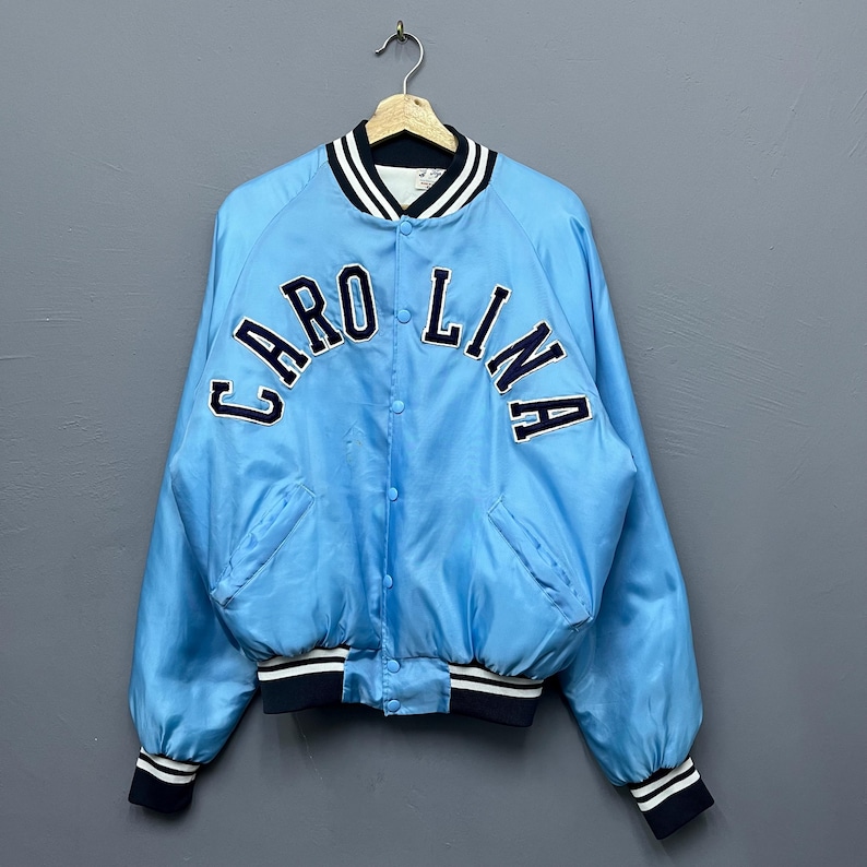 May include: Light blue satin bomber jacket with the word "CAROLINA" arched across the chest in navy blue lettering. The jacket has a navy blue and white striped collar, cuffs, and hem. Features two front pockets and a button-up closure.