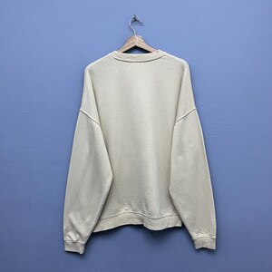 May include: A plain, long-sleeved, cream-colored crewneck sweatshirt made of a solid fabric. The garment features a ribbed collar, ribbed cuffs at the wrists, and a ribbed hemline at the bottom. It is displayed hanging on a wooden hanger.
