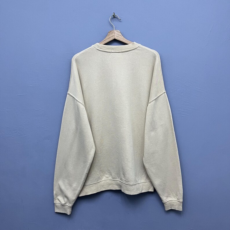 May include: A plain, long-sleeved, cream-colored crewneck sweatshirt made of a solid fabric. The garment features a ribbed collar, ribbed cuffs at the wrists, and a ribbed hemline at the bottom. It is displayed hanging on a wooden hanger.